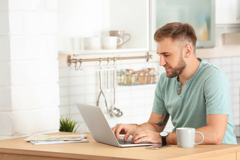 Young handsome man working with laptop at table in kitchen - Photo, Image