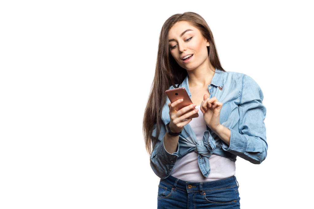 Πορτρέτο της μια μελαχρινή νεαρή γυναίκα κοιτάζοντας το smartphone με έκπληξη έκφραση στο πρόσωπό της ενάντια σε λευκό φόντο, απομονωμένη. Τρόπου ζωής, άνθρωποι και τεχνολογία έννοια - Φωτογραφία, εικόνα