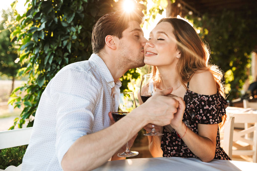 Picture of young loving couple sitting in cafe by dating outdors in park holding glasses of wine drinking kissing. - Photo, Image