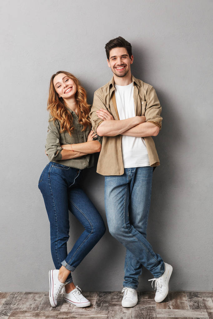 Retrato de larga duración de una alegre pareja joven de pie junto con los brazos cruzados aislados sobre gris
 - Foto, Imagen