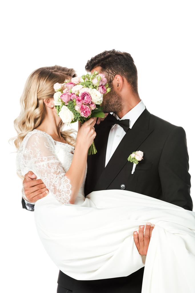 young newlyweds covering faces with bridal bouquet while groom carrying bride isolated on white - Photo, Image