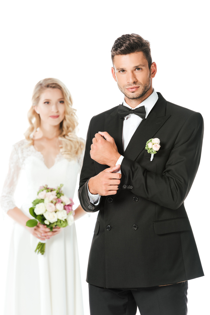 ハンサムな若い新郎カフスボタンをボタンと白で隔離の背景にぼやけている立っている花嫁とカメラ目線 - 写真・画像