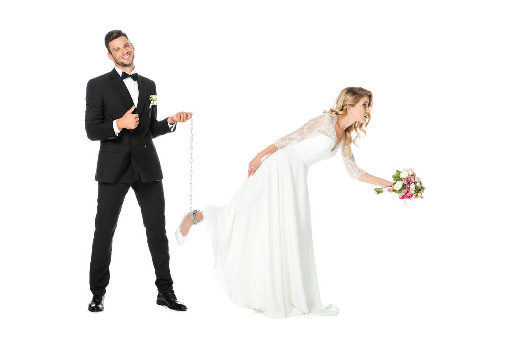 šťastný ženich drží řetěz uvázanou kolem nevěsty nohu a ukazující palec nahoru izolované na bílém - Fotografie, Obrázek