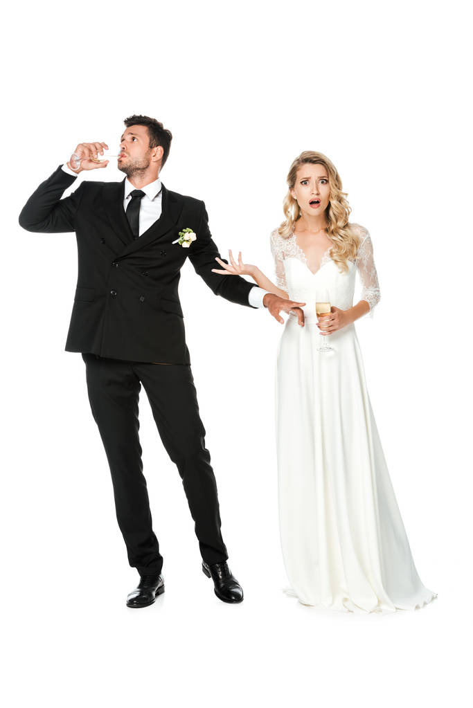 scioccato giovane sposa guardando la fotocamera mentre lo sposo beve champagne isolato su bianco
 - Foto, immagini
