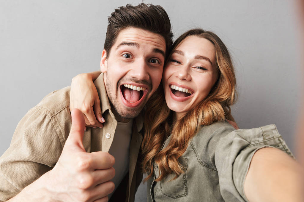 Portrait of an excited young couple hugging while taking a selfie and showing thumbs up isolated over gray background - Photo, Image
