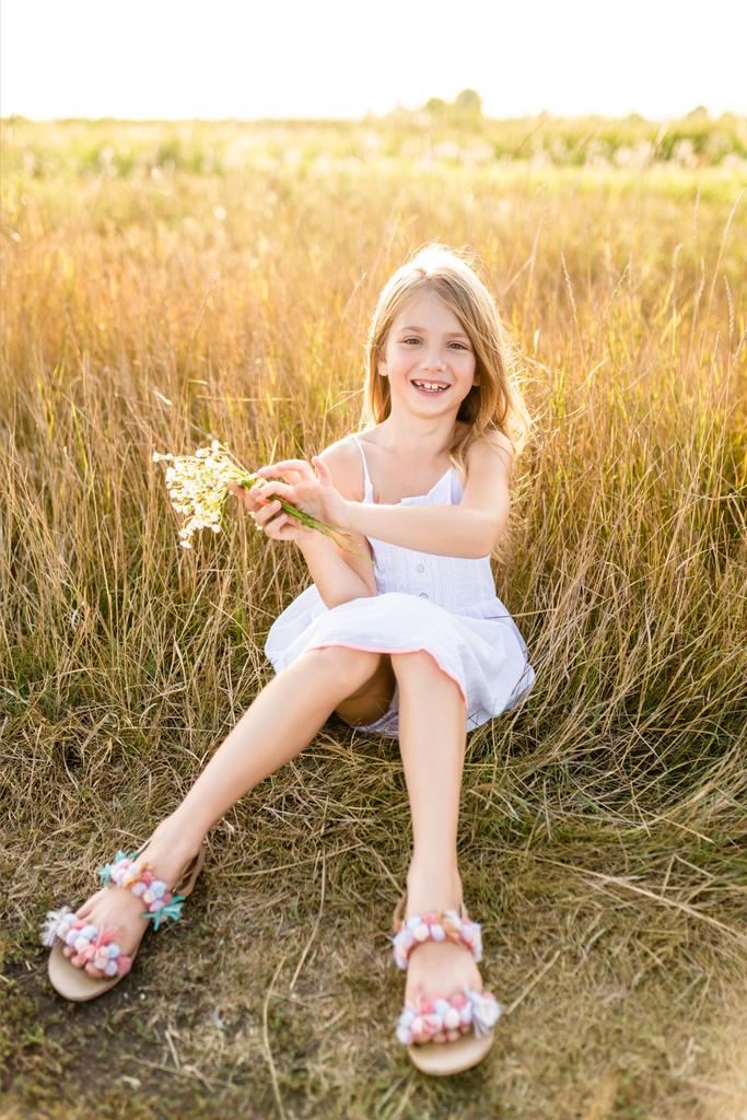 happy little child in white dress with field flowers bouquet sitting in field - Photo, Image