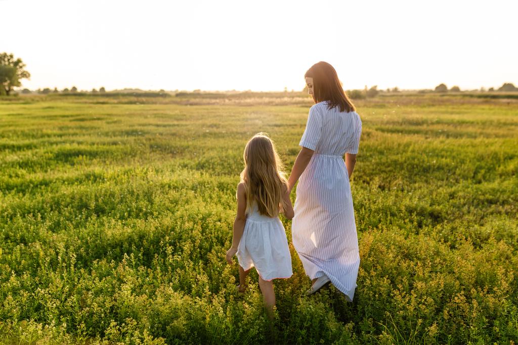 mãe e filha de mãos dadas e caminhando pelo prado verde com o céu por do sol no fundo
 - Foto, Imagem