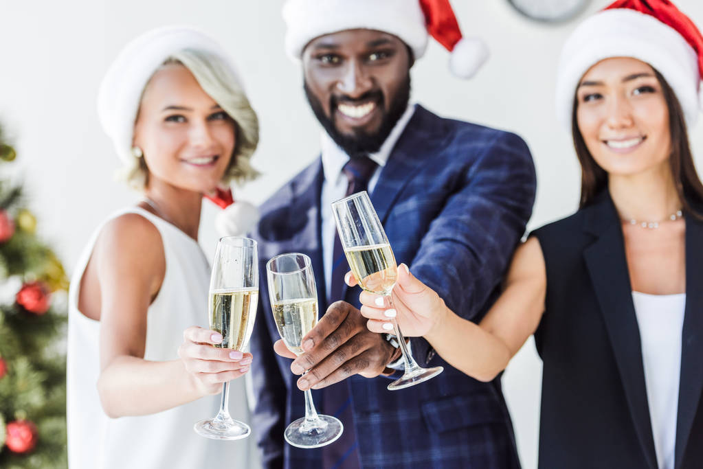 мультиэтнические бизнесмены в шляпах Санта-Клауса звонят с бокалами шампанского в офисе и смотрят в камеру
 - Фото, изображение