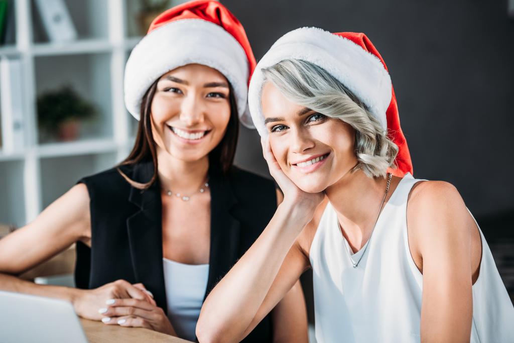 gülümseyen çok kültürlü iş kadınları ofiste kamera bakarak Noel Baba şapkaları  - Fotoğraf, Görsel