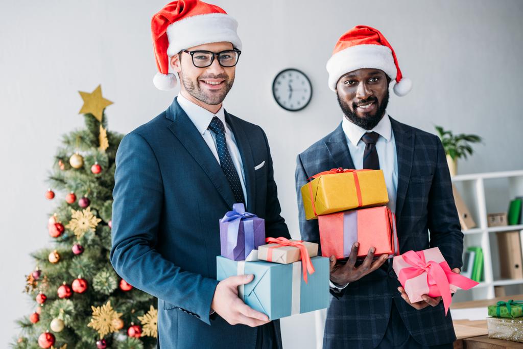 glimlachend multiculturele zakenlieden in santa hoeden holding geschenk dozen in office en de camera kijken - Foto, afbeelding