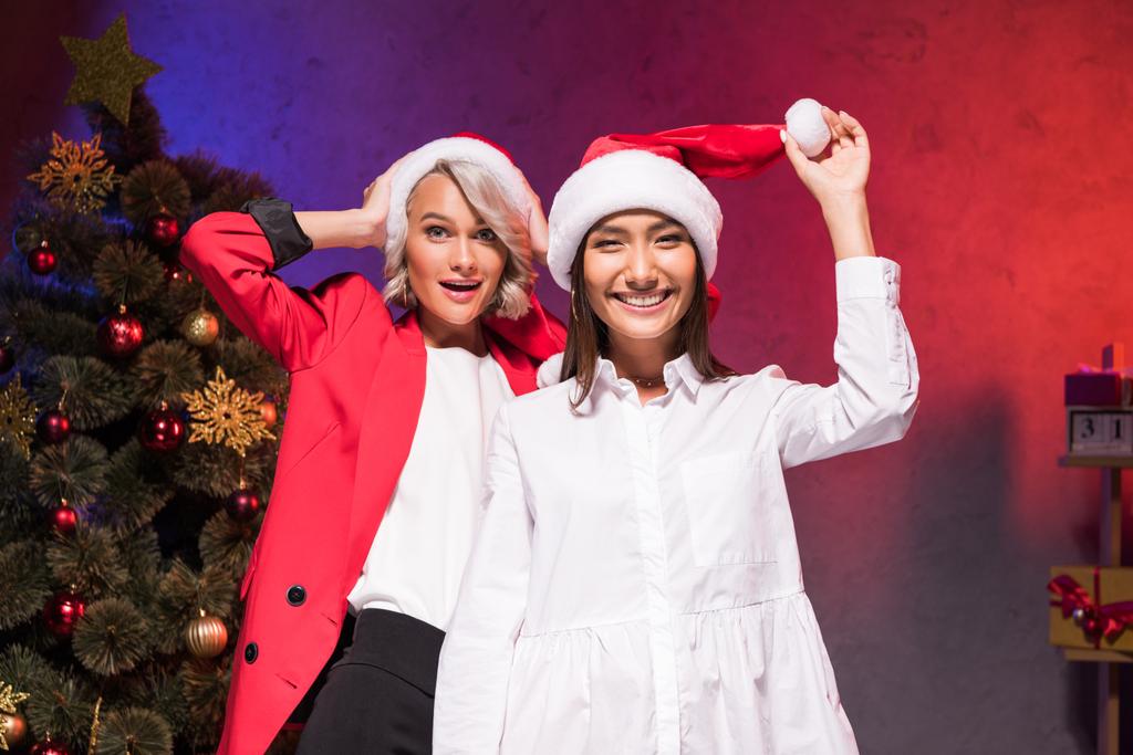 щасливі мультикультурні бізнес-леді тримають капелюхи Санти в офісі на новорічній корпоративній вечірці
 - Фото, зображення
