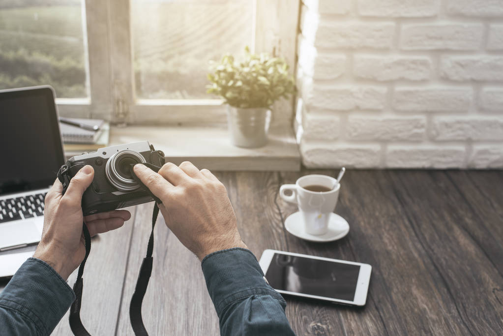 Hipster fotógrafo en casa sentado en el escritorio y la limpieza de su lente de la cámara con un paño, punto de vista de la toma
 - Foto, Imagen