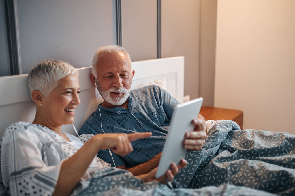 Um casal de idosos se divertindo na cama com um tablet, enquanto também compartilha um fone de ouvido cada
. - Foto, Imagem