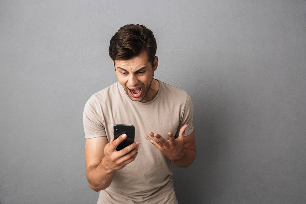 Zirytowany facet 20s w t-shirt krzyczy w gniewie patrząc smartphone na białym tle nad szarym tle - Zdjęcie, obraz
