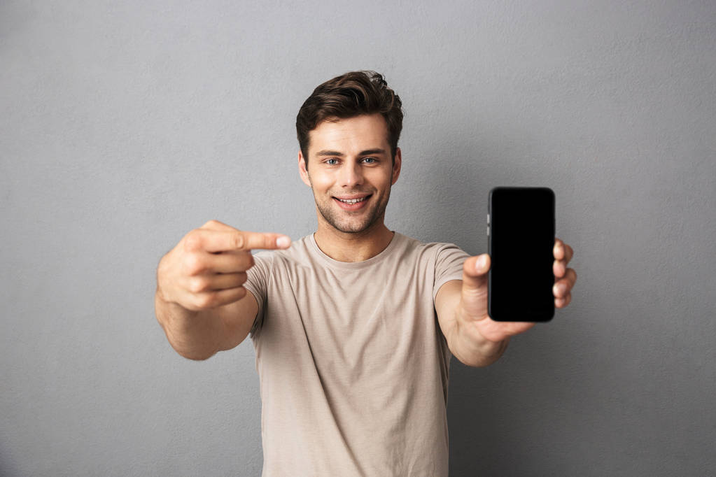 Portret van een vrolijke jongeman in t-shirt over grijze achtergrondkleur, geïsoleerd met leeg scherm mobiele telefoon - Foto, afbeelding