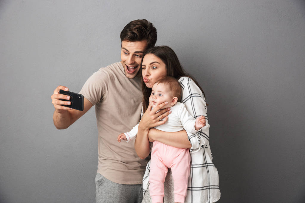 Portrait d'une jeune famille joyeuse avec leur petite fille isolée sur fond gris, prenant un selfie
 - Photo, image