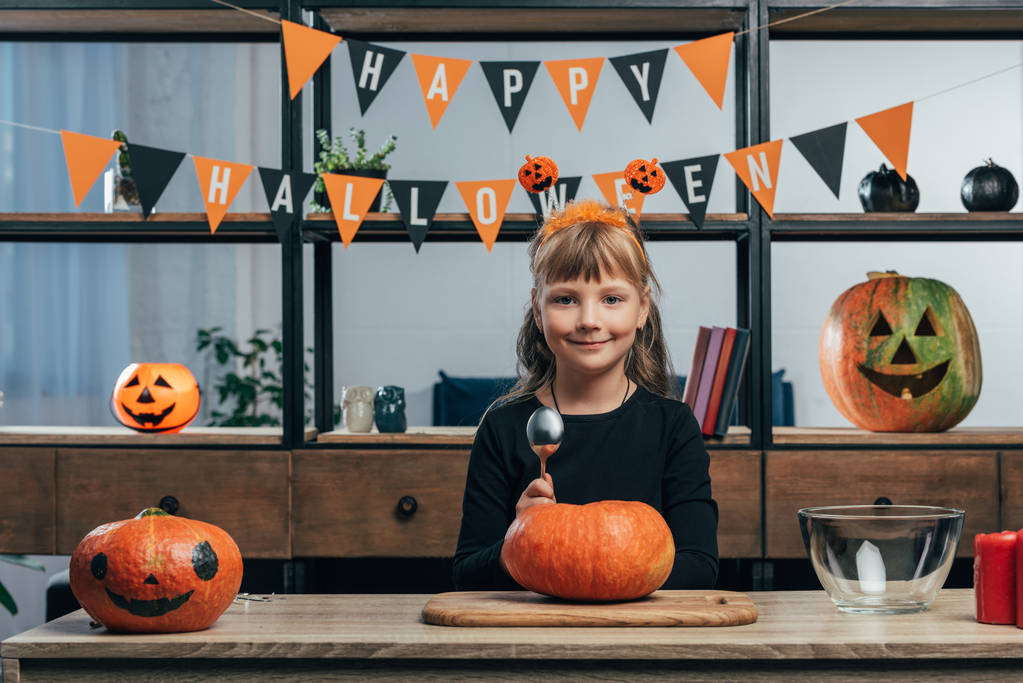 portrait of smiling kid at tabletop with pumpkins and hanging happy halloween flags behind at home - Photo, Image