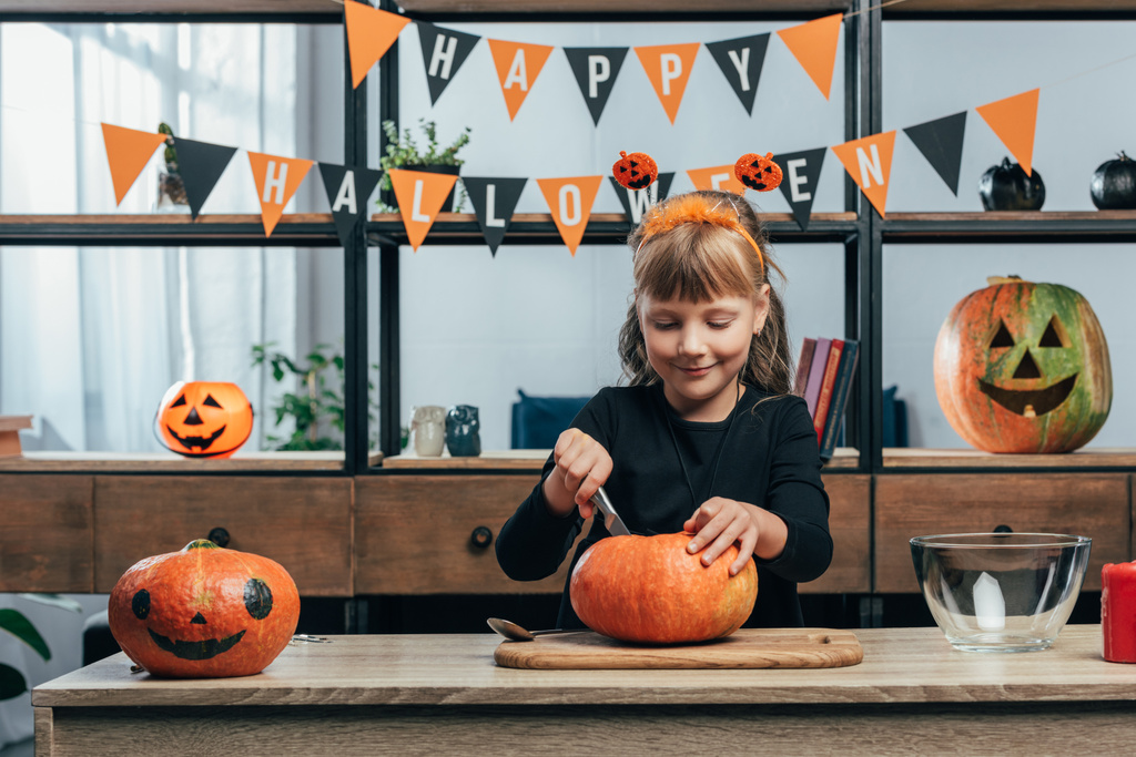 Porträt eines lächelnden Kindes, das zu Hause einen Kürbis schnitzt und fröhliche Halloween-Fahnen aufhängt - Foto, Bild