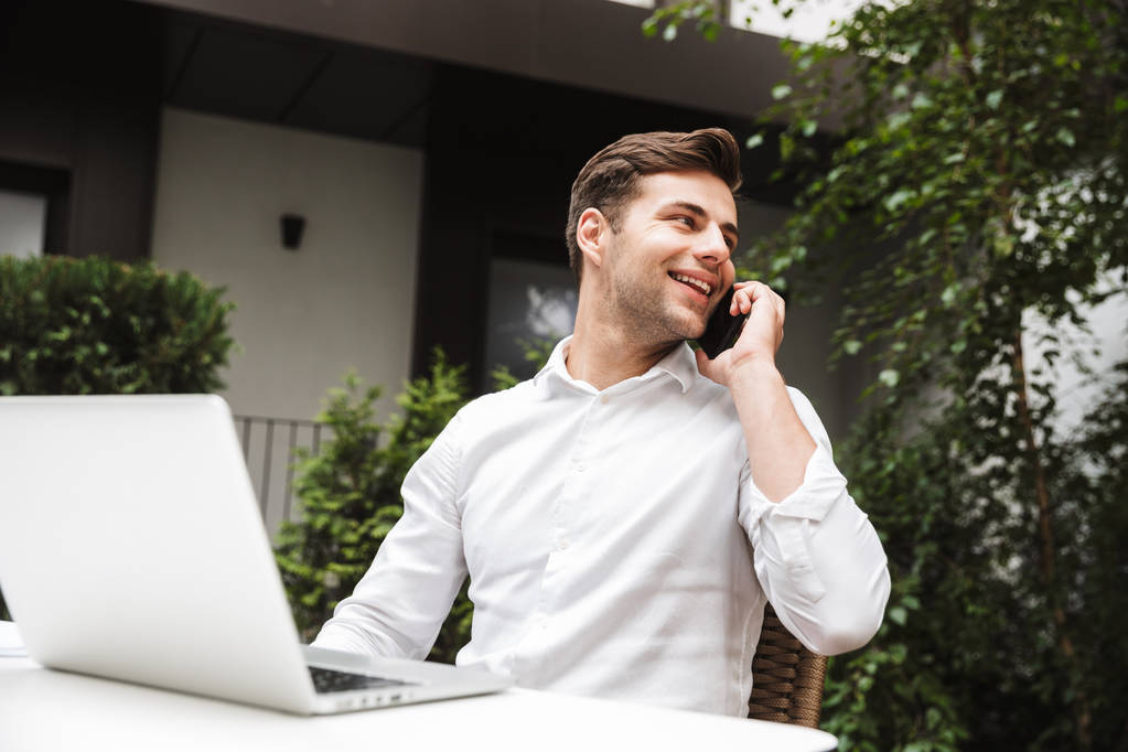 Glimlachend jonge zakenman gekleed in t-shirt praten op mobiele telefoon tijdens het zitten met laptop computer outdoors - Foto, afbeelding