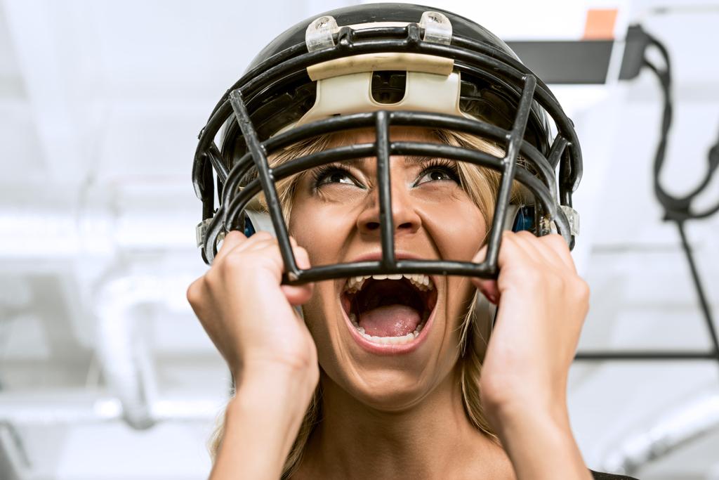 Blick von unten auf eine verrückte junge Frau, die schreit und einen amerikanischen Fußballhelm auf dem Kopf hält - Foto, Bild