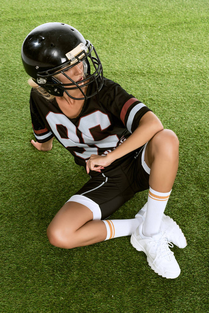 атлетичная молодая женщина в американской футбольной форме сидит на траве и смотрит в сторону
 - Фото, изображение