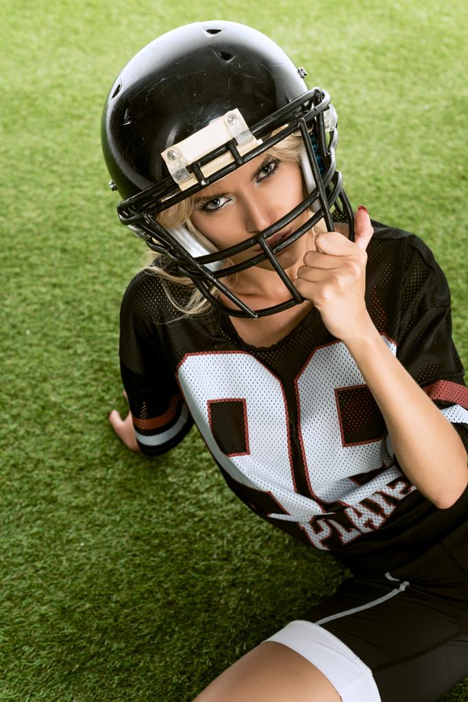 boze jonge vrouw in uniforme zittend van Amerikaans voetbal op gras - Foto, afbeelding