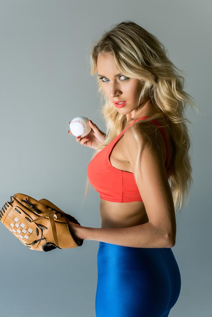 Close-up πορτρέτο της ελκυστική νεαρή γυναίκα σε αθλητικά ενδύματα με Μπέιζμπολ γάντι και μπάλα κοιτάζοντας την κάμερα στο γκρι - Φωτογραφία, εικόνα