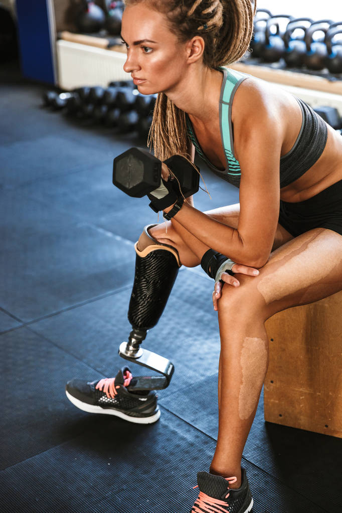 Portrait of feminine handicapped sportswoman wearing prosthesis in tracksuit training and lifting dumbbell in gym - Photo, Image