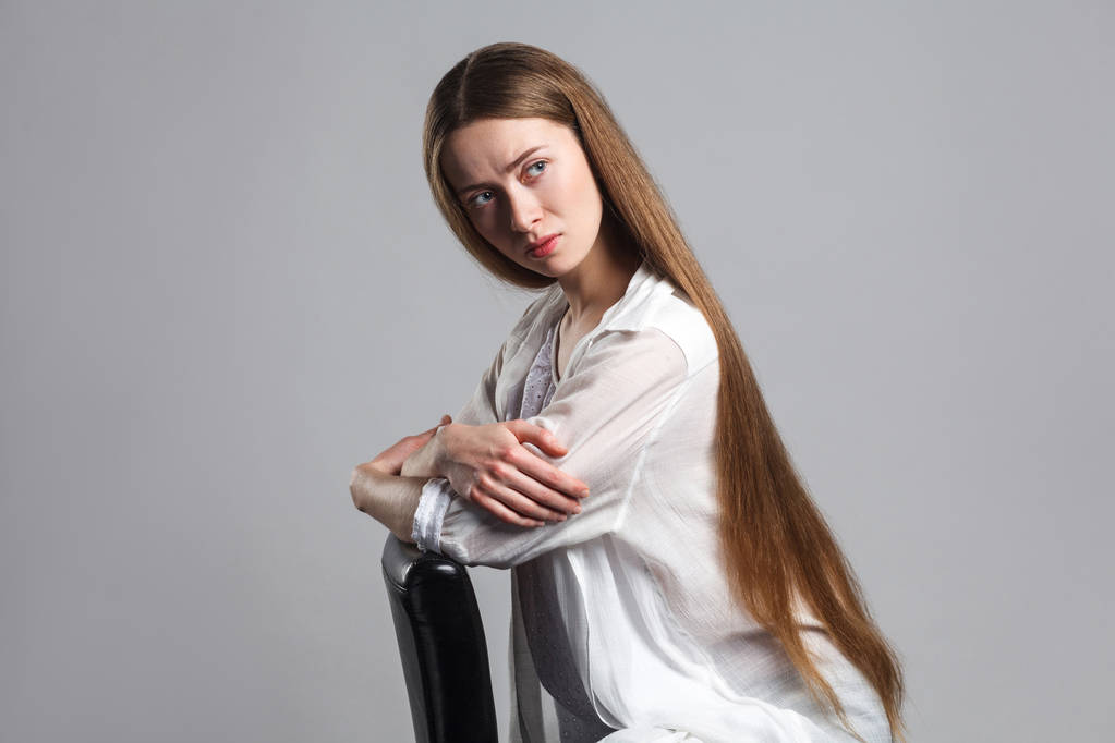 Porträt emotionaler Sorgen junger Model-Schauspieler mit langen braunen Haaren in weißem T-Shirt sitzt auf schwarzem Stuhl mit gekreuzten Händen und blickt mit ernstem verwirrten Gesicht auf grauem Hintergrund weg - Foto, Bild