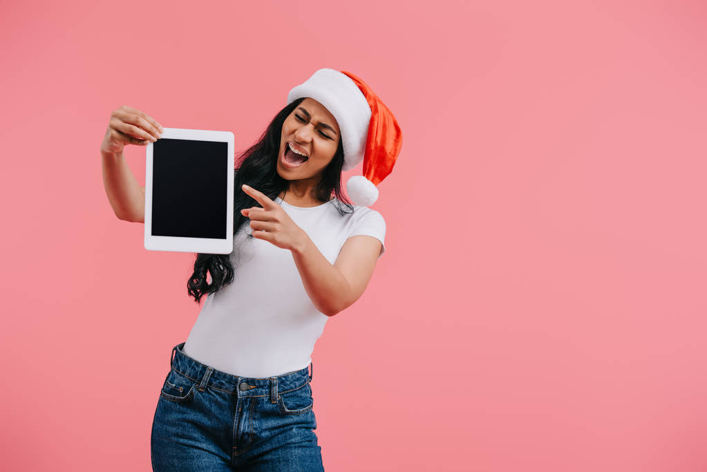 Porträt einer emotionalen afrikanisch-amerikanischen Frau mit Weihnachtsmann-Hut, die auf Tablet mit leerem Bildschirm zeigt, isoliert auf rosa  - Foto, Bild