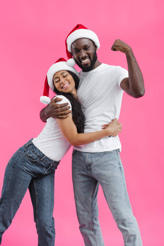 attractive african american woman in christmas hat embracing boyfriend while he showing muscles on hand isolated on pink background  - Photo, Image