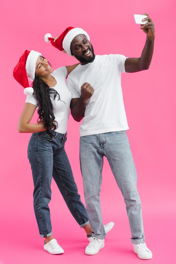 giovane uomo afroamericano in cappello di Natale mostrando pugno e prendendo selfie su smartphone con fidanzata su sfondo rosa
 - Foto, immagini