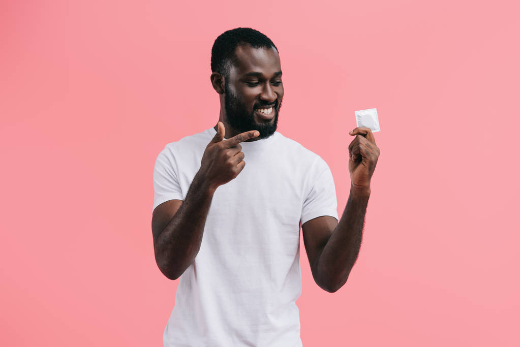 hymyilevä nuori afrikkalainen amerikkalainen mies osoittaa kondomi eristetty vaaleanpunainen tausta
  - Valokuva, kuva