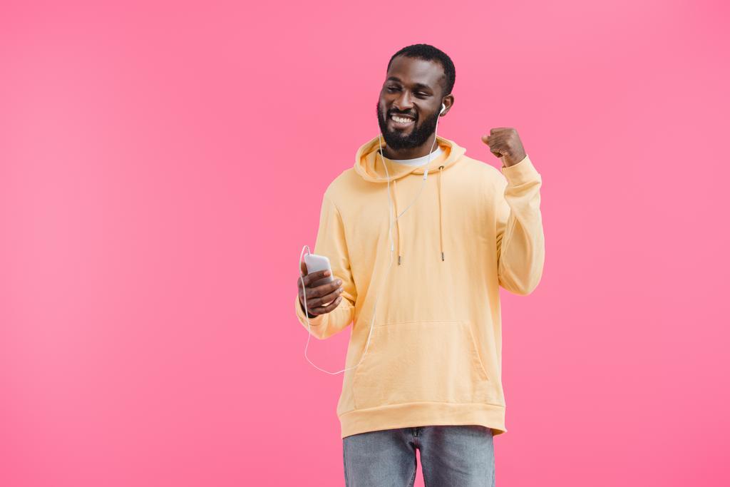 happy african american man in earphones gesturing by hand listening music with smartphone isolated on pink background - Photo, Image