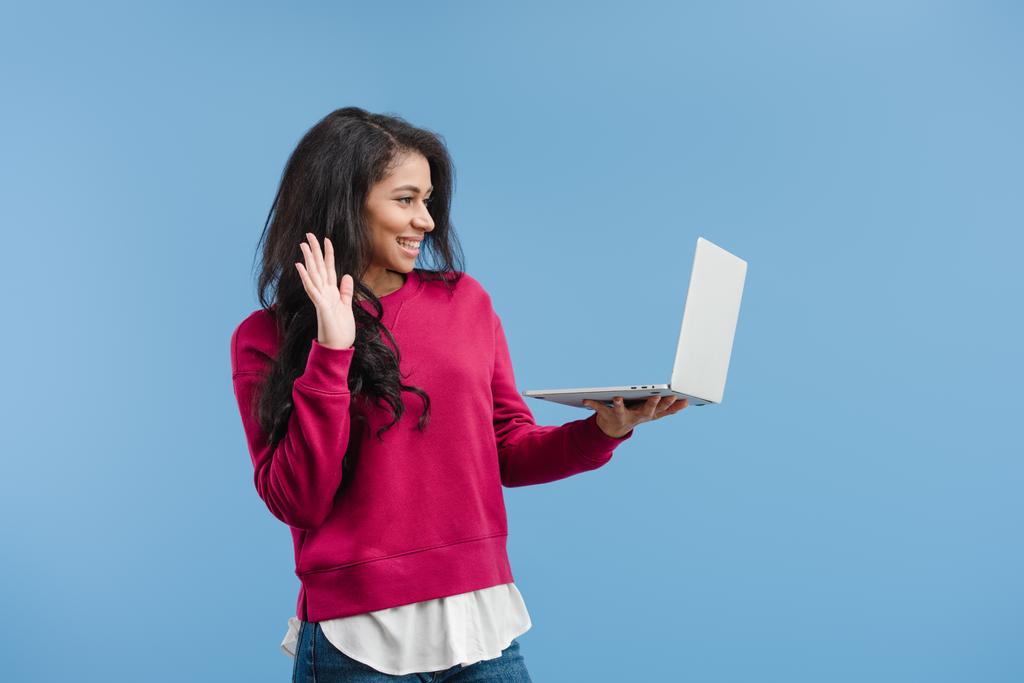 vista lateral da mulher americana africana feliz acenando à mão e ter chamada de vídeo no laptop isolado no fundo azul
  - Foto, Imagem