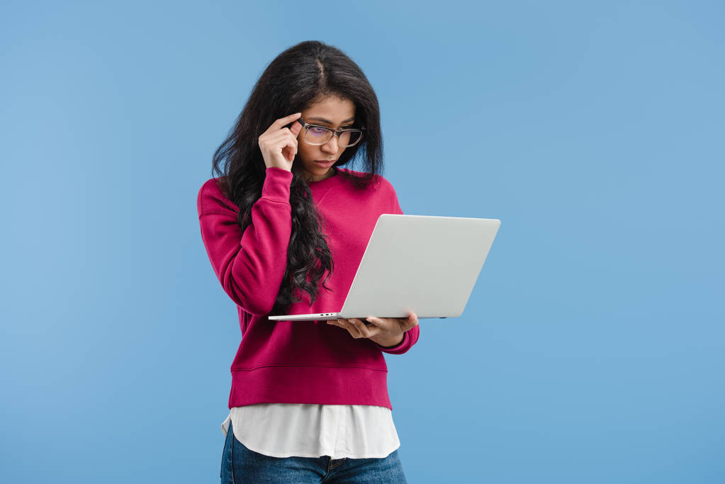 serious young african american woman adjusting eyeglasses and looking at laptop isolated on blue background  - Photo, Image