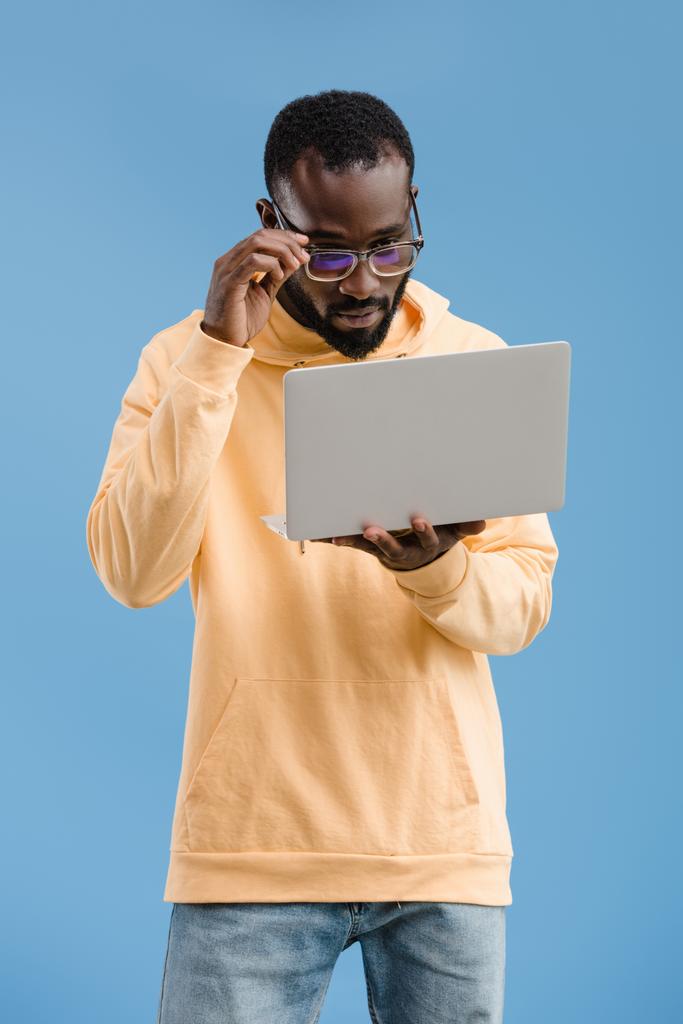 young african american man adjusting eyeglasses and looking at laptop isolated on blue background - Photo, Image