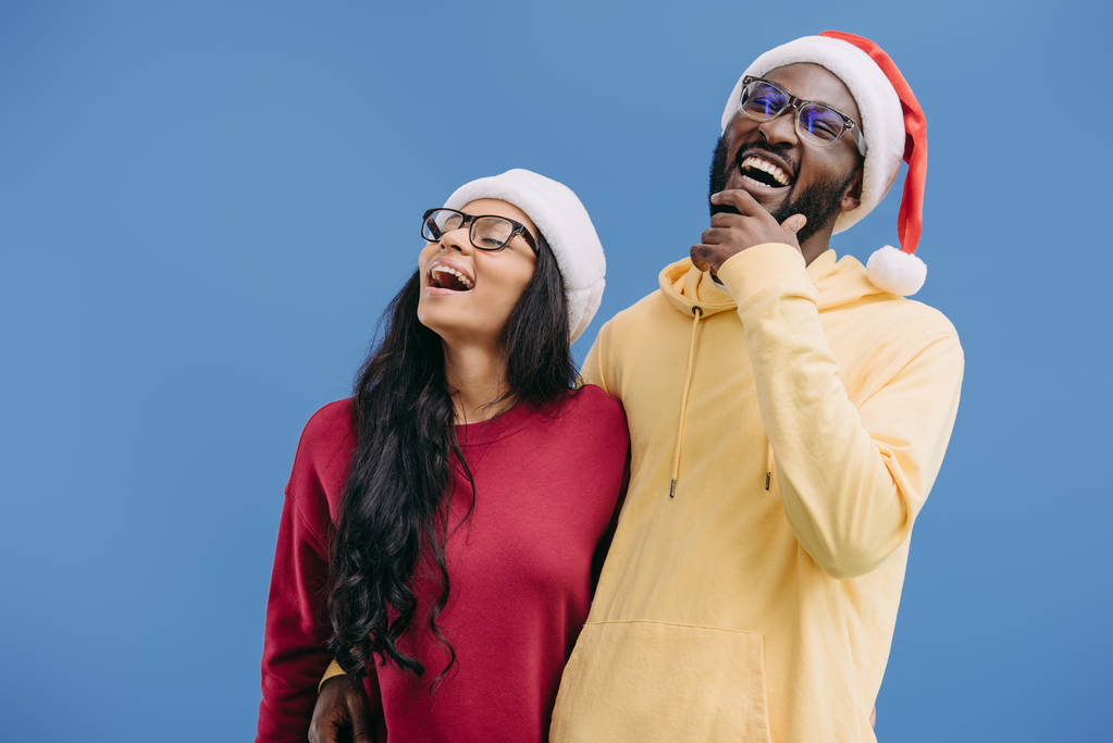 γελώντας αφρικανική αμερικανική ζευγάρι στο Χριστούγεννα καπέλα και γυαλιά που απομονώνονται σε μπλε φόντο  - Φωτογραφία, εικόνα