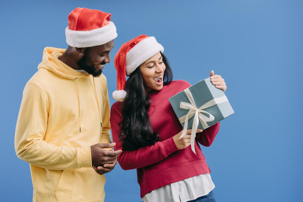 aufgeregte afrikanisch-amerikanische Frau mit Weihnachtsmütze und Geschenkbox, während ihr Freund in der Nähe isoliert auf blauem Hintergrund steht  - Foto, Bild