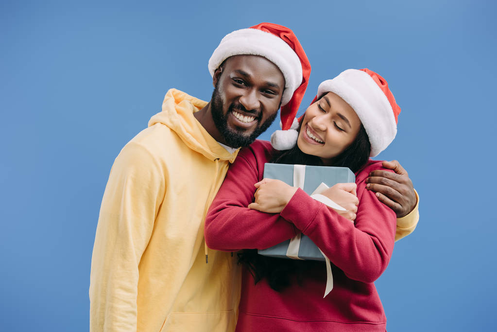 счастливый африканский американец в рождественской шляпе обнимает подругу с подарочной коробкой изолированы на синем фоне
  - Фото, изображение