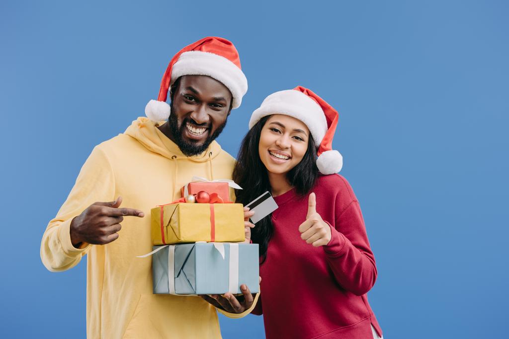 afro americano uomo in cappello di Natale che punta a scatole regalo mentre la sua ragazza in possesso di carta di credito e facendo pollice in su gesto isolato su sfondo blu
 - Foto, immagini