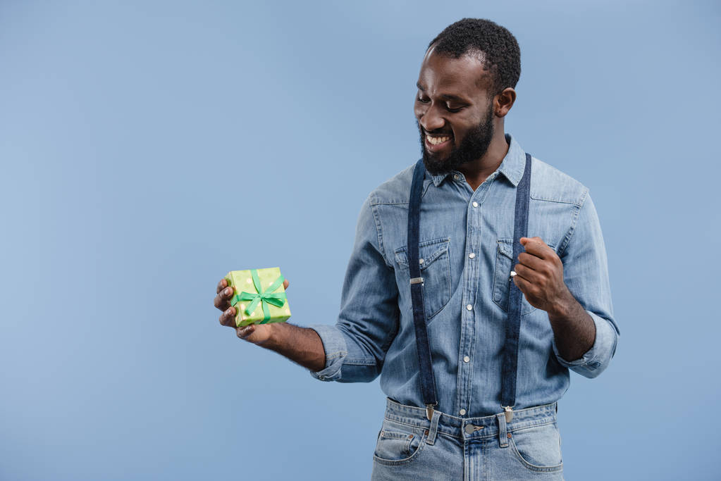 feliz joven afroamericano hombre con caja de regalo envuelto por cinta gesto a mano aislado sobre fondo azul
 - Foto, Imagen