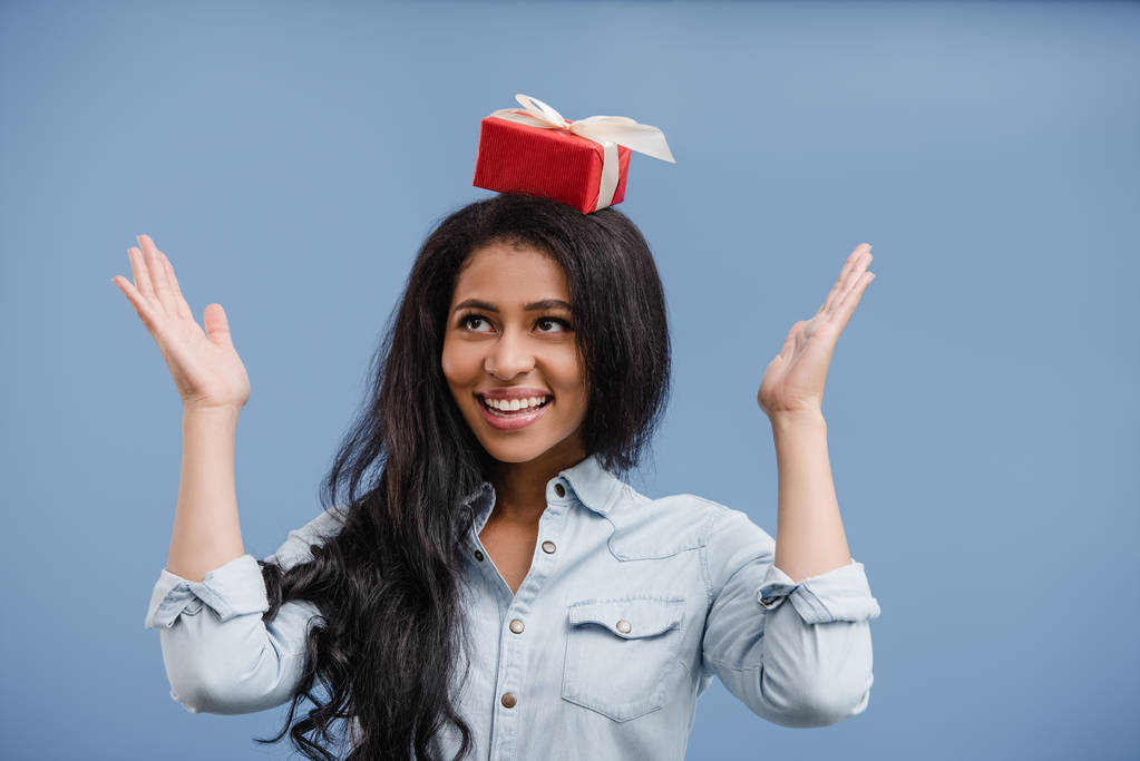 feliz atraente menina americana africana segurando caixa de presente vermelho na cabeça isolada no azul
 - Foto, Imagem