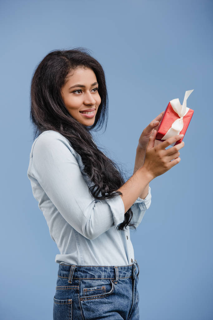 vrolijke aantrekkelijke Afro-Amerikaanse meisje houdt van de rode luxe-geschenketui en op zoek weg geïsoleerd op blauw - Foto, afbeelding