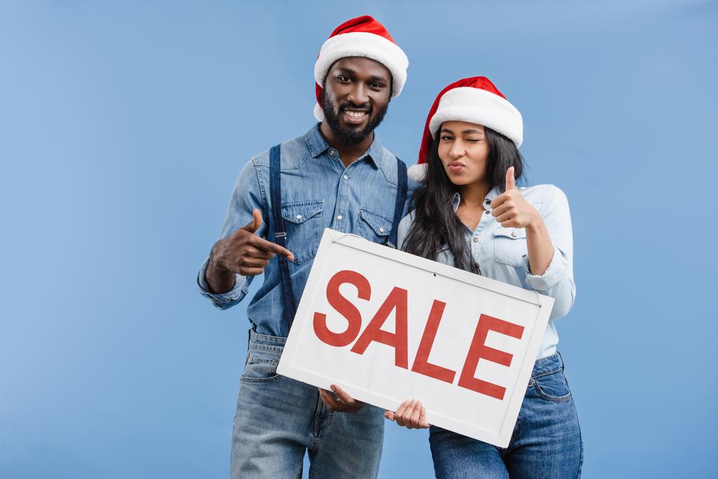 coppia afro-americana in cappelli di Babbo Natale indicando in vendita segno isolato su blu
 - Foto, immagini