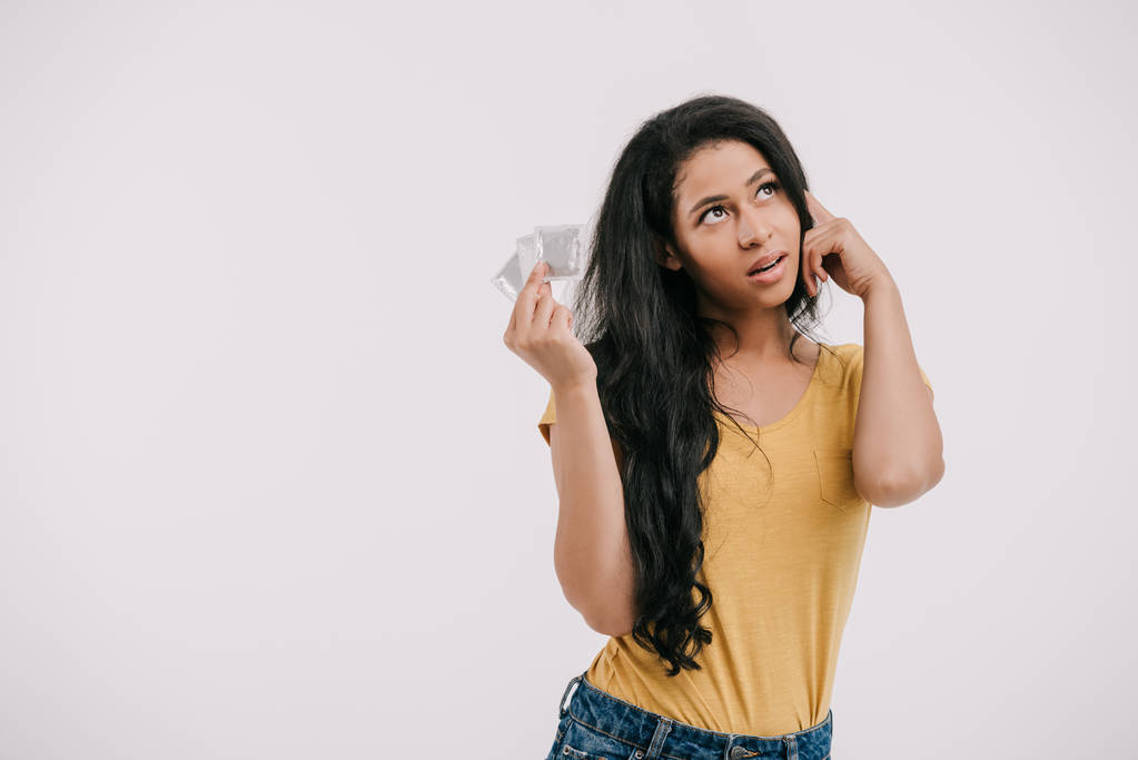 nadenkend aantrekkelijke Afro-Amerikaanse meisje houdt van latex condooms en opzoeken van geïsoleerde op wit - Foto, afbeelding