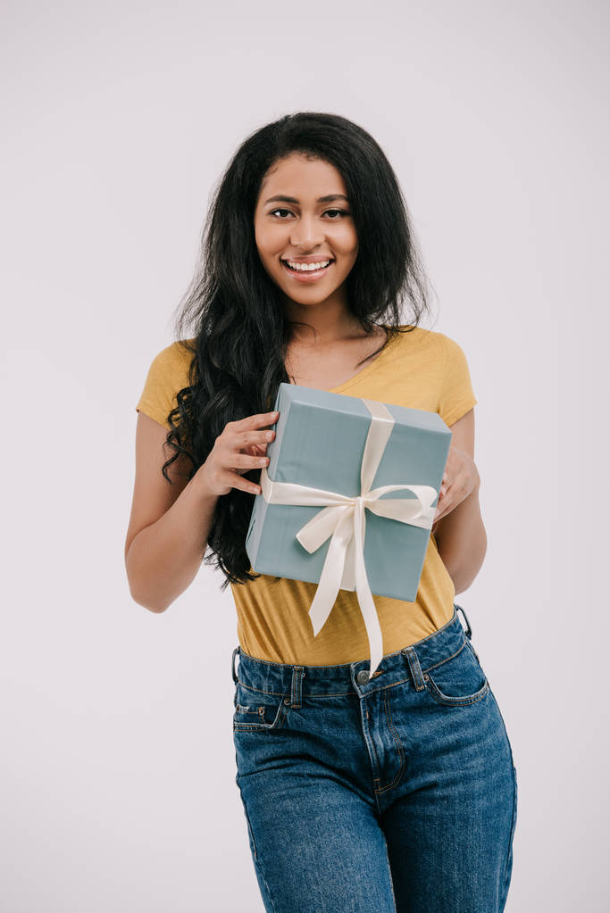 sorridente ragazza afroamericana in possesso di una scatola regalo e guardando la fotocamera isolata su bianco
  - Foto, immagini