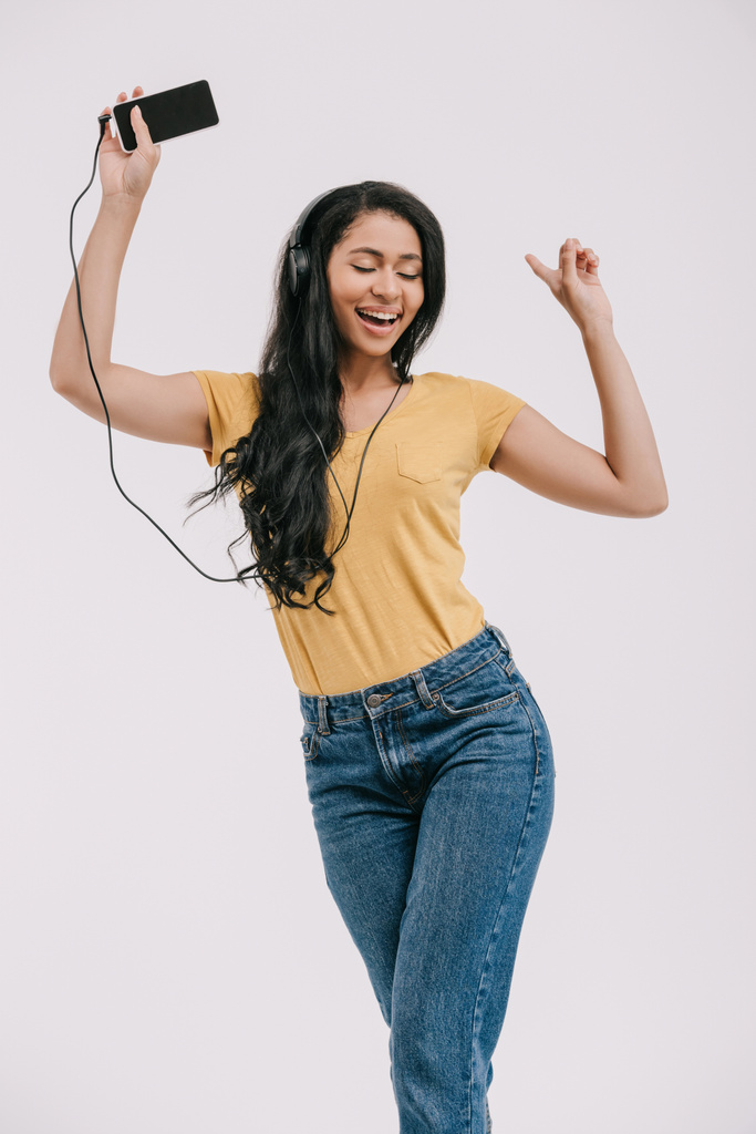 atractiva chica afroamericana bailando mientras escucha música con teléfono inteligente y auriculares aislados en blanco
 - Foto, imagen