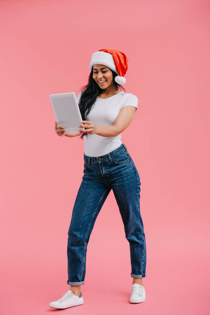 uśmiechający się atrakcyjna dziewczyna Afryki amerykański w santa hat patrząc na tablecie na różowym tle - Zdjęcie, obraz