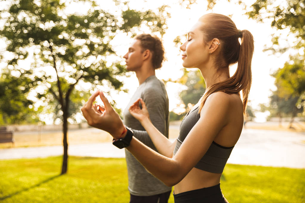 Wizerunek zrelaksowany sportowy Para mężczyzna i kobieta lat 20 w dresy, trzymając palce razem do medytacji podczas treningu albo robi joga w parku - Zdjęcie, obraz